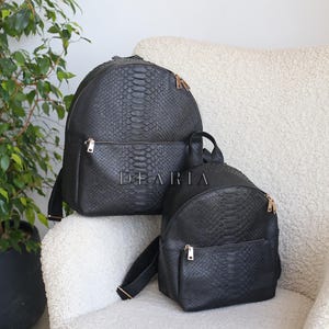 Könnte beinhalten: Zwei schwarze Rucksäcke aus Kunstleder mit Schlangenmuster in verschiedenen Größen. Der größere Rucksack hat eine Vordertasche und goldfarbene Reißverschlüsse. Der kleinere Rucksack hat ebenfalls eine Vordertasche und goldfarbene Reißverschlüsse. Auf dem größeren Rucksack steht "DEARIA".