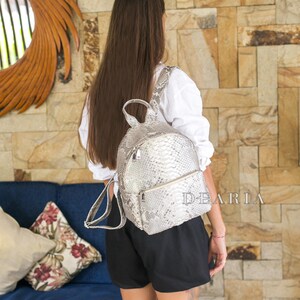 May include: A silver snakeskin-patterned backpack with a zipped front pocket and adjustable straps. The brand name "DEARIA" is visible. The backpack is worn with a white shirt and black shorts. The bag is a stylish accessory.