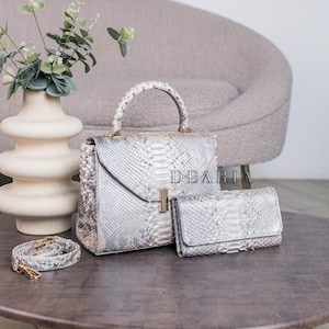 Peut inclure: Ensemble de sacs à main et portefeuille en cuir à motif serpent. L'ensemble comprend un sac à poignée supérieure, un portefeuille et une bandoulière. Les sacs sont gris clair et blancs avec des accessoires dorés. Le mot "DEARIA" est imprimé.