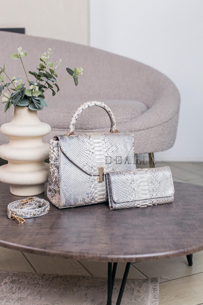 K&ouml;nnte beinhalten: Ein Set aus Handtaschen und einer Geldb&ouml;rse mit Schlangenmuster. Das Set beinhaltet eine Tasche mit oberem Griff, eine Geldb&ouml;rse und einen Schulterriemen. Die Taschen sind hellgrau und wei&szlig; mit goldfarbener Hardware. Das Wort "DEARIA" ist auf der Tasche aufgedruckt.