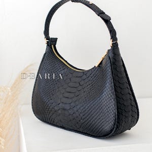 Pu&ograve; includere: Una borsa a mano nera in pelle di serpente con una cerniera dorata e un manico superiore.
