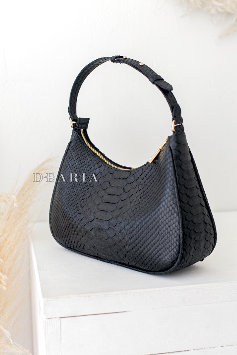 Pu&ograve; includere: Una borsa a mano nera in pelle di serpente con una cerniera dorata e un manico superiore.