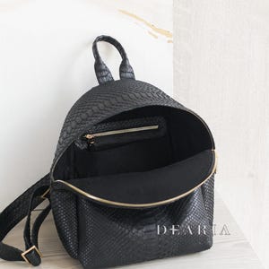 May include: Black backpack with a textured, snakeskin-like pattern. The bag is open, revealing a black interior and a small zipped pocket. The zip and hardware are gold-coloured. The word "DEARIA" is printed on the side.