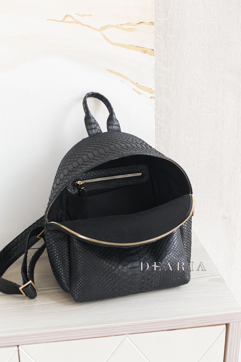 May include: Black backpack with a textured, snakeskin-like pattern. The bag is open, revealing a black interior and a small zipped pocket. The zip and hardware are gold-coloured. The word "DEARIA" is printed on the side.