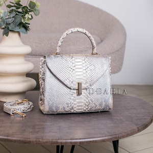 K&ouml;nnte beinhalten: Eine silber-graue Handtasche aus Schlangenhaut mit einem oberen Griff und einem abnehmbaren Schulterriemen. Die Tasche hat eine rechteckige Form mit einem Klappenverschluss und einem goldfarbenen Verschluss. Das Wort "DEARIA" ist auf der Tasche aufgedruckt.