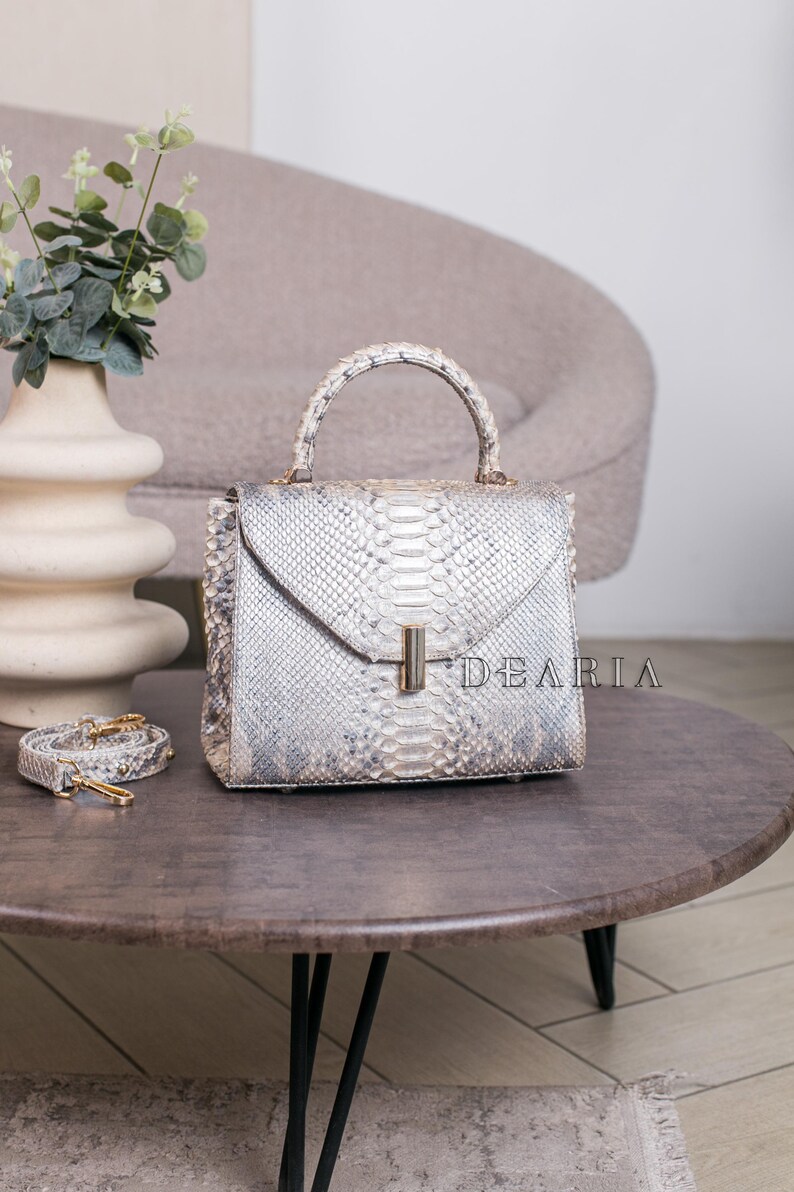 K&ouml;nnte beinhalten: Eine silber-graue Handtasche aus Schlangenhaut mit einem oberen Griff und einem abnehmbaren Schulterriemen. Die Tasche hat eine rechteckige Form mit einem Klappenverschluss und einem goldfarbenen Verschluss. Das Wort "DEARIA" ist auf der Tasche aufgedruckt.