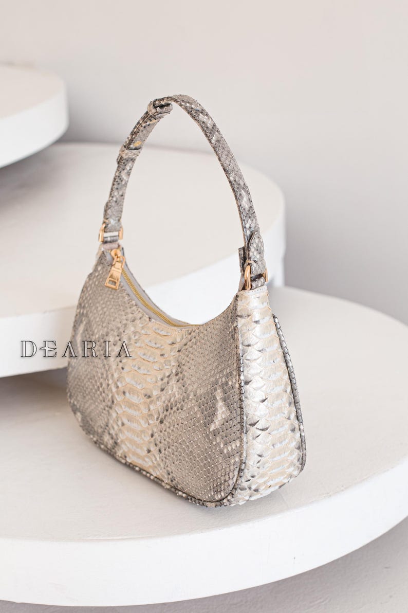 Pu&ograve; includere: Una piccola borsa a mano in pelle di serpente grigio chiaro con un manico corto e curvo. La borsa ha una chiusura a zip e un motivo testurizzato sottile.