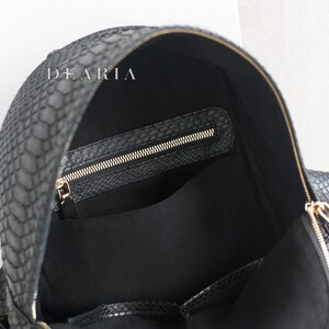 May include: Black backpack with a snakeskin texture and gold-coloured zips. The interior is black with a zipped pocket and a large open pocket. The brand name "DEARIA" is visible on the top.