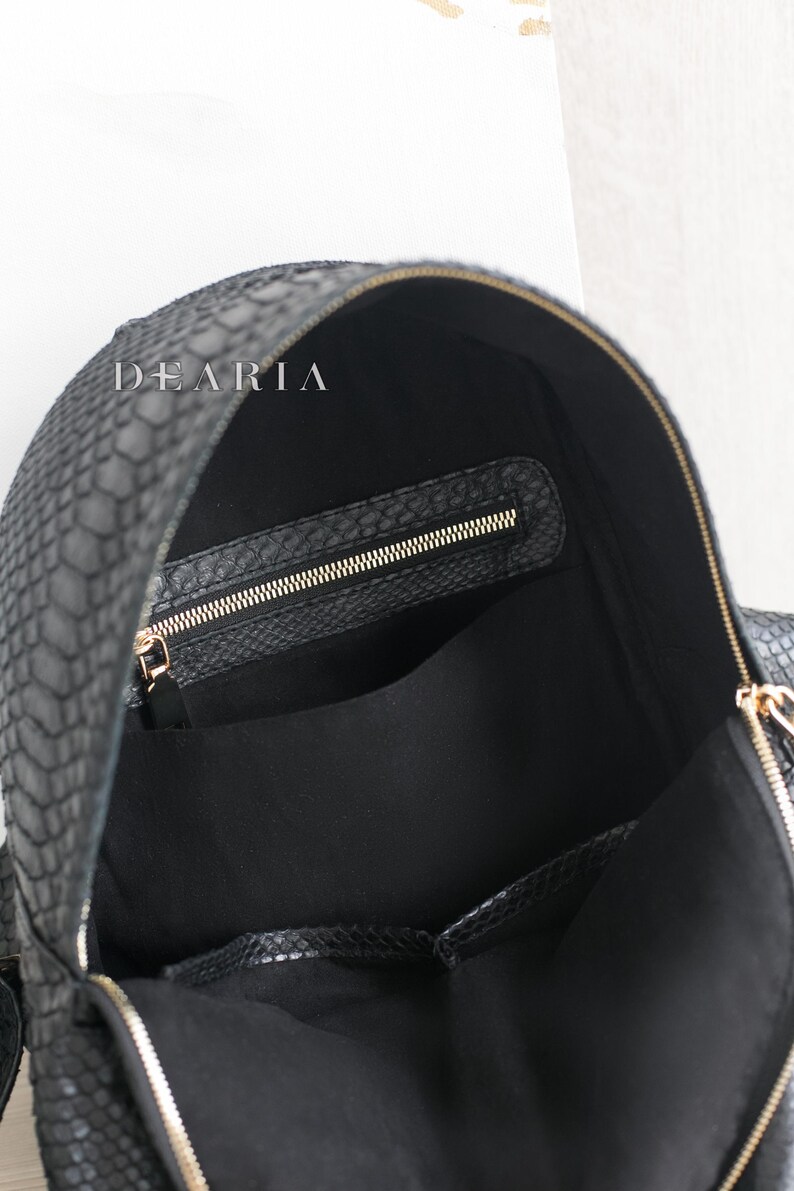 May include: Black backpack with a snakeskin texture and gold-coloured zips. The interior is black with a zipped pocket and a large open pocket. The brand name "DEARIA" is visible on the top.