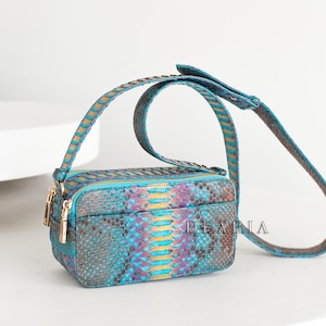 Könnte beinhalten: Eine rechteckige Umhängetasche mit einem lebendigen Schlangenmuster in Türkis-, Lila- und Goldtönen. Die Tasche hat einen goldenen Reißverschluss und einen passenden Riemen. Der Markenname "DE ARIA" ist auf der Vorderseite sichtbar.