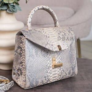 K&ouml;nnte beinhalten: Eine strukturierte Handtasche mit Schlangenmuster in Grau- und Wei&szlig;t&ouml;nen. Die Tasche hat einen oberen Griff, einen Klappverschluss und goldfarbene Hardware. Das Wort "DEARIA" ist auf der Tasche aufgedruckt.