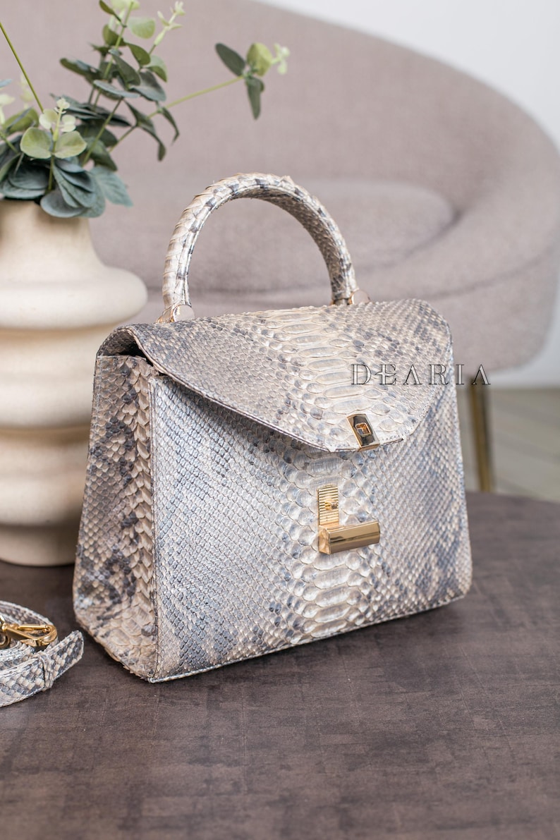 K&ouml;nnte beinhalten: Eine strukturierte Handtasche mit Schlangenmuster in Grau- und Wei&szlig;t&ouml;nen. Die Tasche hat einen oberen Griff, einen Klappverschluss und goldfarbene Hardware. Das Wort "DEARIA" ist auf der Tasche aufgedruckt.