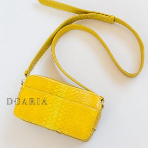 May include: A bright yellow rectangular crossbody bag with a matching adjustable strap. The bag features a textured, snake-like pattern and a zippered closure. The word "DEARIA" is printed on the front of the bag.