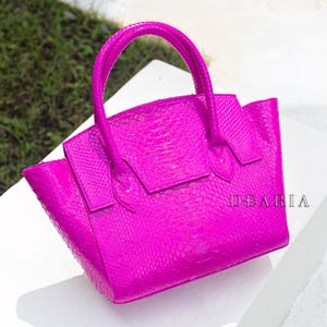 May include: A bright fuchsia handbag with a textured, snakeskin-like pattern. The bag has a structured design with a top handle and a flap closure. The word "DEARIA" is visible on the side of the bag.