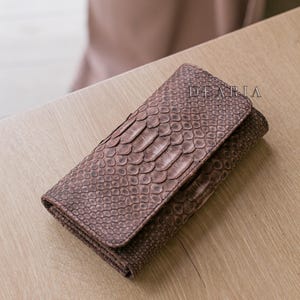 Może przedstawiać: Brązowy skórzany portfel o fakturze skóry gada. Portfel jest prostokątny z klapką. Na górze portfela wydrukowano napis "DEARIA".