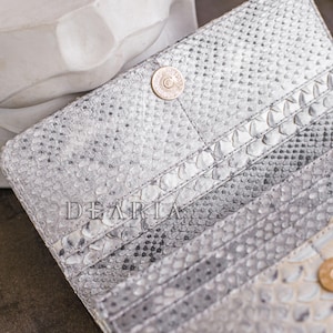 Puede incluir: Cartera abierta, texturizada, plateada y blanca, con cierre de bot&oacute;n a presi&oacute;n. La cartera tiene m&uacute;ltiples ranuras para tarjetas y la palabra "DEARIA" est&aacute; impresa en la parte delantera. La cartera est&aacute; hecha de un material similar a la piel de serpiente.