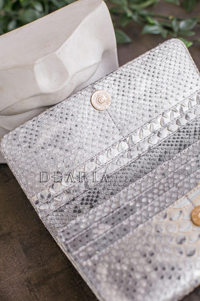 Puede incluir: Cartera abierta, texturizada, plateada y blanca, con cierre de bot&oacute;n a presi&oacute;n. La cartera tiene m&uacute;ltiples ranuras para tarjetas y la palabra "DEARIA" est&aacute; impresa en la parte delantera. La cartera est&aacute; hecha de un material similar a la piel de serpiente.