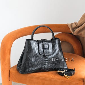 Könnte beinhalten: Eine schwarze Ledertasche mit Schlangenhautmuster und einem goldenen Schnallenverschluss. Die Tasche liegt auf einem orangefarbenen Stuhl mit einem passenden Riemen.