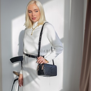 May include: A woman in a white dress with a black rectangular crossbody bag. The bag has a textured pattern, gold zips and hardware. The text "DEARIA" is visible. She wears a gold necklace and a black belt. The bag's strap is black.