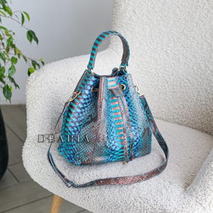 May include: A blue and brown snakeskin-patterned bucket bag with a handle and a shoulder strap. The bag is sitting on a white chair. The bag has a drawstring closure and gold-colored hardware. The word "DEARIA" is printed on the bag.