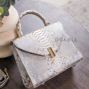 K&ouml;nnte beinhalten: Handtasche mit wei&szlig;em und grauem Schlangenmuster, mit goldfarbenem Verschluss und Griff. Die Tasche hat eine strukturierte Form und einen Klappenverschluss. Das Wort "DEARIA" ist auf der Tasche aufgedruckt.