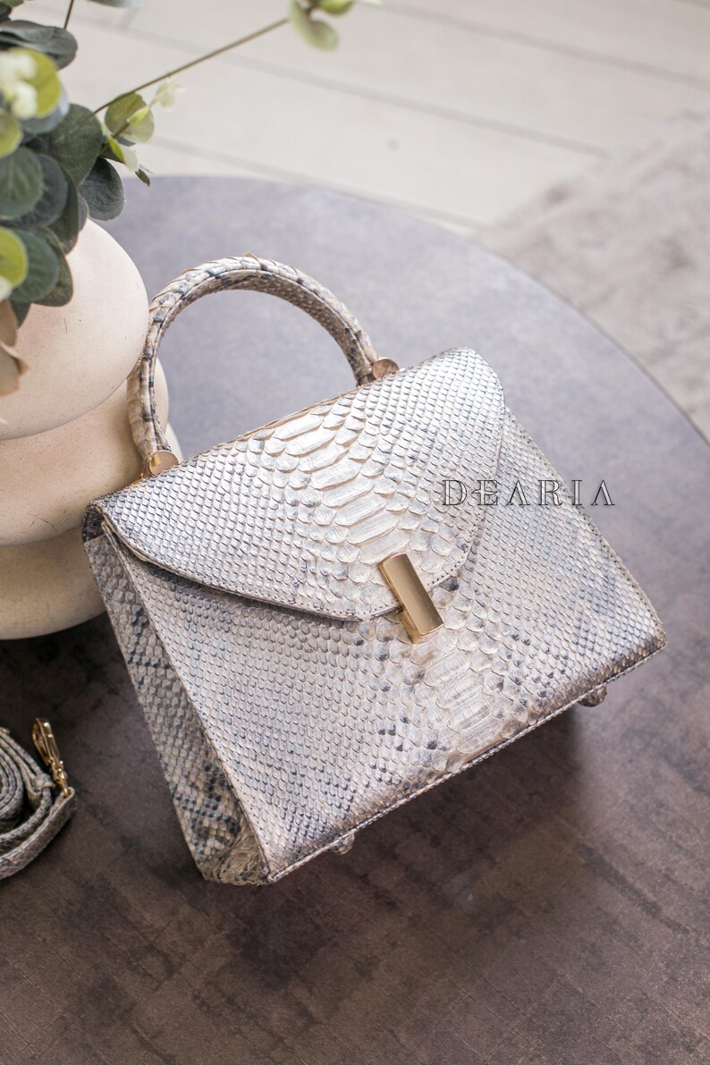 K&ouml;nnte beinhalten: Handtasche mit wei&szlig;em und grauem Schlangenmuster, mit goldfarbenem Verschluss und Griff. Die Tasche hat eine strukturierte Form und einen Klappenverschluss. Das Wort "DEARIA" ist auf der Tasche aufgedruckt.