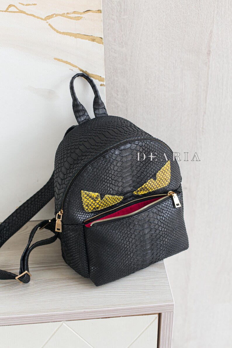 May include: Black backpack with a reptile skin texture, featuring yellow and black eye-shaped accents. The bag has a zippered front pocket with a gold zip and a red interior. The brand name "DEARIA" is printed on the bag.