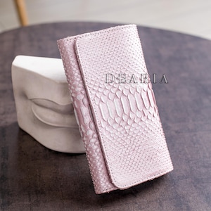 May include: A light pink leather wallet with a snakeskin pattern. The wallet has a flap closure and the word "DEARIA" is printed on the front. The wallet is resting on a dark surface.