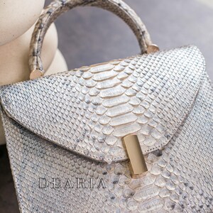 K&ouml;nnte beinhalten: Silber-wei&szlig; Handtasche aus Schlangenleder mit gebogenem Griff und goldfarbenem Verschluss. Das Wort "DEARIA" ist auf der Vorderseite eingepr&auml;gt. Die Tasche hat einen Klappverschluss und ein strukturiertes Design.