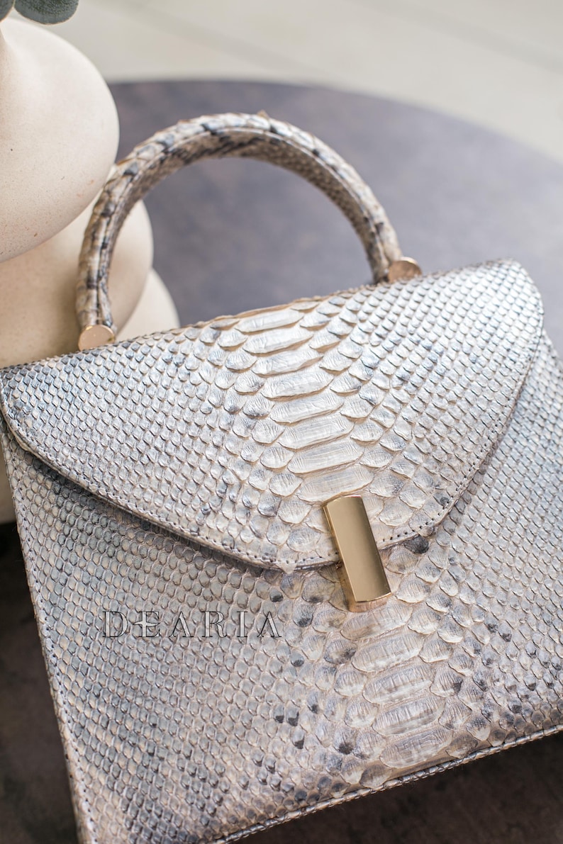 K&ouml;nnte beinhalten: Silber-wei&szlig; Handtasche aus Schlangenleder mit gebogenem Griff und goldfarbenem Verschluss. Das Wort "DEARIA" ist auf der Vorderseite eingepr&auml;gt. Die Tasche hat einen Klappverschluss und ein strukturiertes Design.