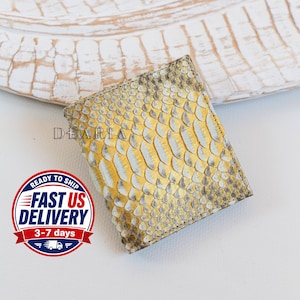 May include: A square wallet featuring a textured snakeskin pattern in shades of yellow, grey, and white. The word DEARIA is printed in black on the front surface of the wallet. The wallet has visible stitching along the edges and a flat, compact design.