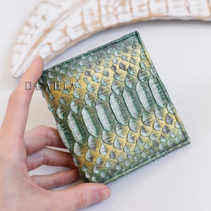 May include: A square wallet with a textured, scaly pattern in shades of green and gold. The wallet is held by a hand, with the word "DEARIA" visible on the side. The background is a blurred white surface and a decorative wooden element.