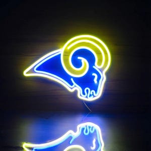 Puede incluir: Letrero de neón con forma de cabeza de carnero, con cabeza azul y cuerno amarillo. La luz de neón se refleja en una superficie oscura, creando un efecto de espejo. El letrero es un objeto decorativo.