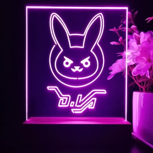 以下が含まれることがあります： 黒い背景にウサギのロゴと「D.Va」の文字が入ったネオンピンクのLEDライト。木製の台座に取り付けられています。画像には、ピンクの花が入った花瓶も含まれており、明るい雰囲気を作り出しています。