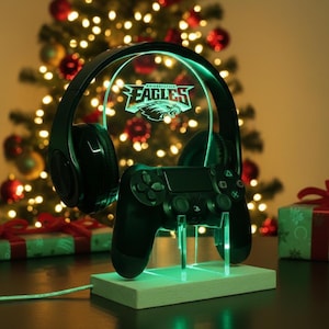 May include: A black gaming headset and controller are displayed on a lighted stand. The stand features the Philadelphia Eagles logo and a green glow. The background includes a Christmas tree with lights and wrapped gifts.