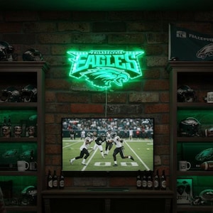 May include: A neon green Philadelphia Eagles sign is mounted on a brick wall above a television. The TV displays a football game. Shelves on either side hold Eagles-themed merchandise, including helmets and mugs. A team flag is visible.