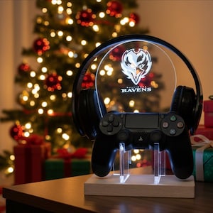 Puede incluir: Un mando de videojuegos negro y unos auriculares descansan sobre una base de madera con un soporte de acrílico transparente. Un panel circular iluminado muestra el logotipo de los Ravens de Baltimore. El fondo presenta un árbol de Navidad con adornos rojos y luces.