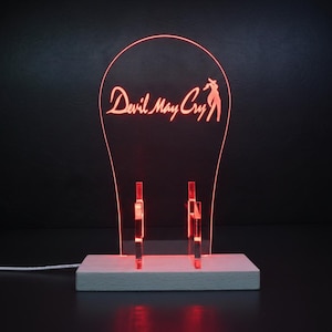 May include: A clear acrylic display stand with a red LED light effect, featuring the text Devil May Cry and a silhouette figure etched into the surface. The stand is mounted on a rectangular wooden base with a white power cord extending to the left.