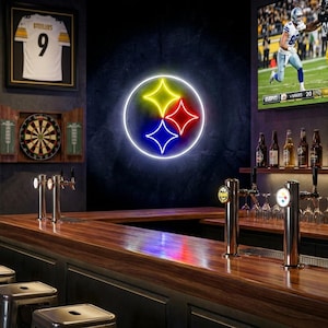 May include: A bar with a neon sign of the Pittsburgh Steelers logo, featuring yellow, red, and blue stars within a white circle. Framed jerseys and a dartboard are visible. A TV displays a football game.