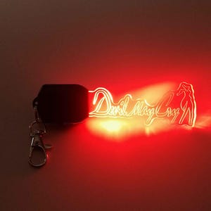 May include: A glowing keychain featuring the text "Devil May Cry" in a stylized font. The keychain has a silhouette of a character and emits a bright red light. It includes a black base and a silver clasp.
