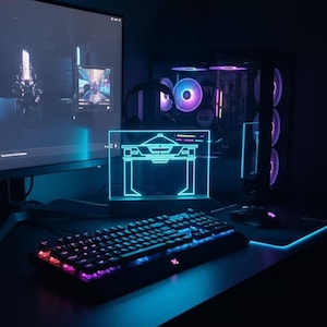 May include: A gaming setup with a backlit keyboard, monitor, and desktop computer. The keyboard has multi-coloured keys. A neon blue acrylic desk accessory is in the foreground. The scene is illuminated with blue and purple lighting.