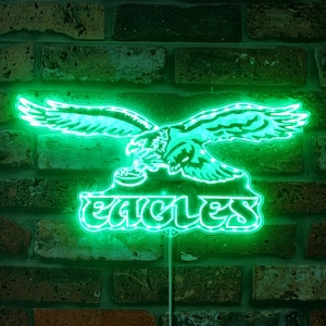 May include: Green neon sign with an eagle design and the word "Eagles" underneath. The sign is lit up and has a white cord.