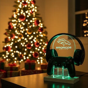 Peut inclure: Un support de casque de jeu éclairé avec le logo des Broncos de Denver et une manette. Le support est illuminé de lumières vertes et repose sur une base en bois. Un sapin de Noël décoré est en arrière-plan.
