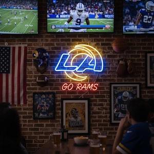 Puede incluir: Una pared de ladrillo decorada con recuerdos deportivos, incluyendo un letrero de neón con las letras "LA" y las palabras "GO RAMS". La pared también presenta carteles de equipos enmarcados, un balón de fútbol y una bandera estadounidense.