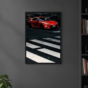 May include: A framed wall art print featuring a bright red sports car parked at a pedestrian crosswalk. The car is angled toward the right, showcasing its sleek front bumper, headlights, and a license plate. The scene is set on dark asphalt.