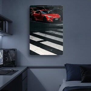 May include: A rectangular wall art print featuring a bright red sports car stopped at a white-striped pedestrian crosswalk on a dark asphalt road. The car is angled toward the right, with visible headlights and a license plate in a city setting.