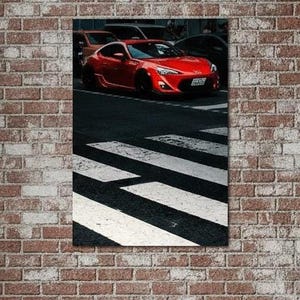 以下が含まれることがあります： 横断歩道に停車している鮮やかな赤色のスポーツカーを描いた長方形のウォールアートプリント。車は暗いアスファルトの上に配置されており、横断歩道の白い縞模様が画像の前面に向かって伸びています。