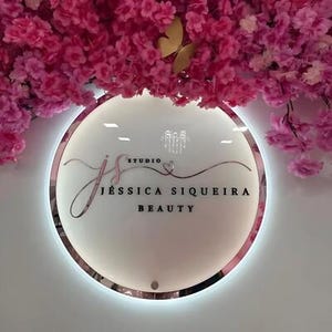 Może przedstawiać: Okrągły, podświetlany znak z napisem "JS Studio JESSICA SIQUEIRA BEAUTY". Znak ma r&oacute;żową obw&oacute;dkę i jest umieszczony na białej ścianie. R&oacute;żowe kwiaty i złoty motyl znajdują się nad znakiem.
