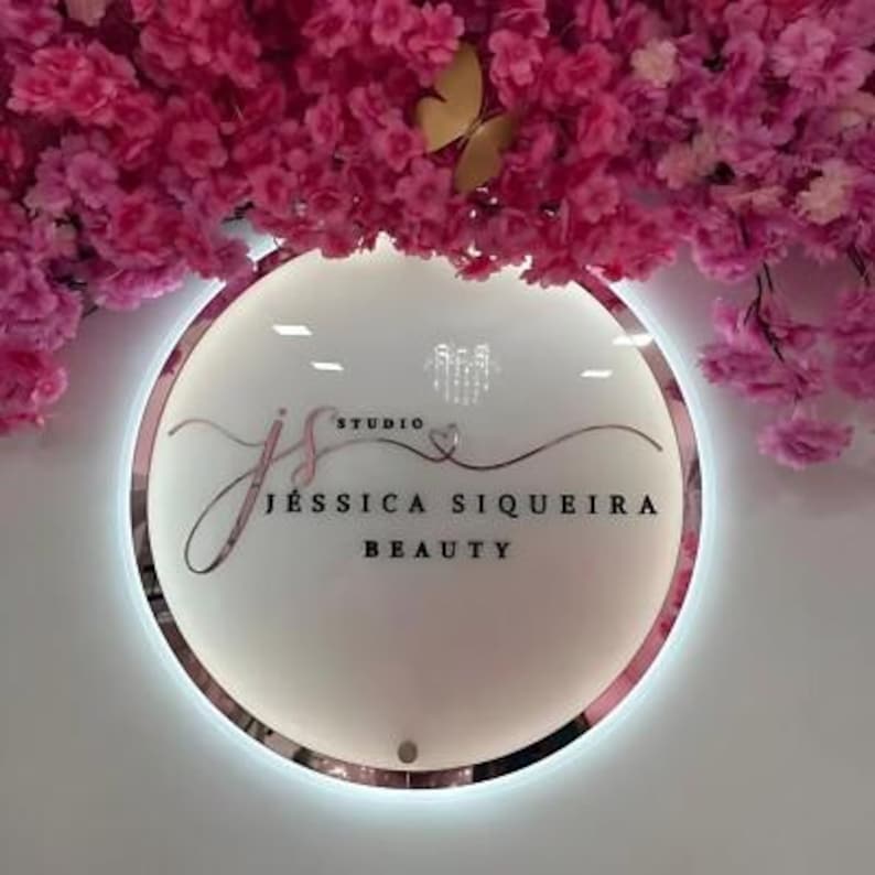 Może przedstawiać: Okrągły, podświetlany znak z napisem "JS Studio JESSICA SIQUEIRA BEAUTY". Znak ma r&oacute;żową obw&oacute;dkę i jest umieszczony na białej ścianie. R&oacute;żowe kwiaty i złoty motyl znajdują się nad znakiem.