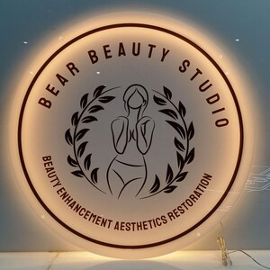Może przedstawiać: Okrągły, podświetlany szyld dla Bear Beauty Studio. Znak ma białe tło z czarnym tekstem i rysunkiem kobiety. Tekst brzmi "BEAR BEAUTY STUDIO" i "BEAUTY ENHANCEMENT AESTHETICS RESTORATION".
